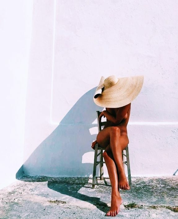 Rear view of a relaxed woman sitting on a wooden chair in Sahra.Nko swimwear, wearing a large straw hat against a white wall, symbolizing tan maintenance