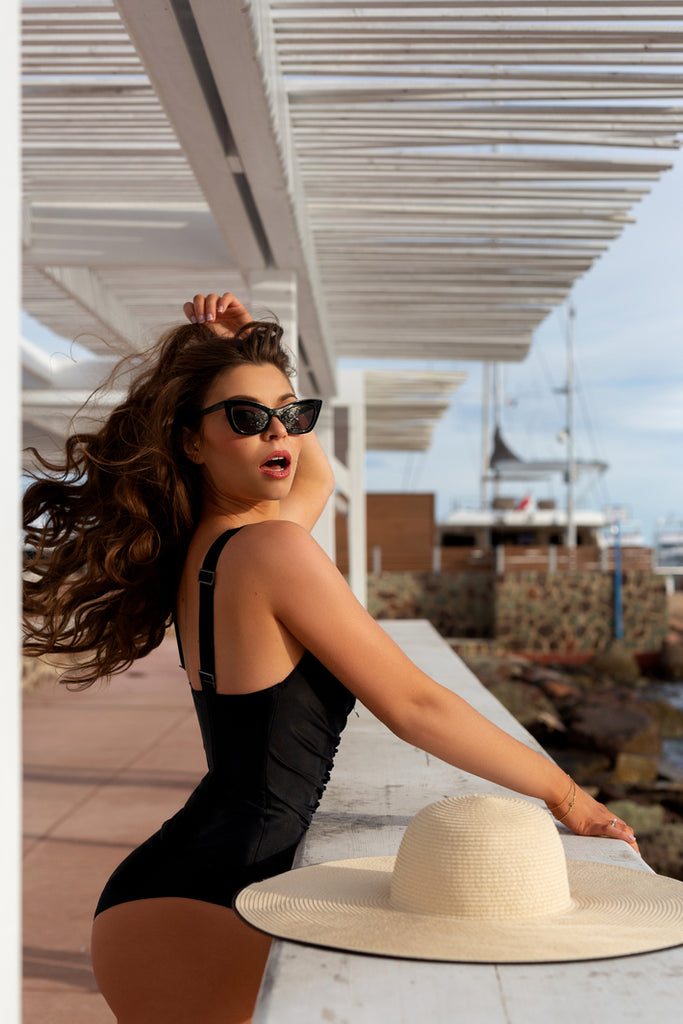 Elegant woman in Cannes wearing Sahra.Nko's black corset swimsuit, confidently posing on a pier with sunglasses and a straw hat.